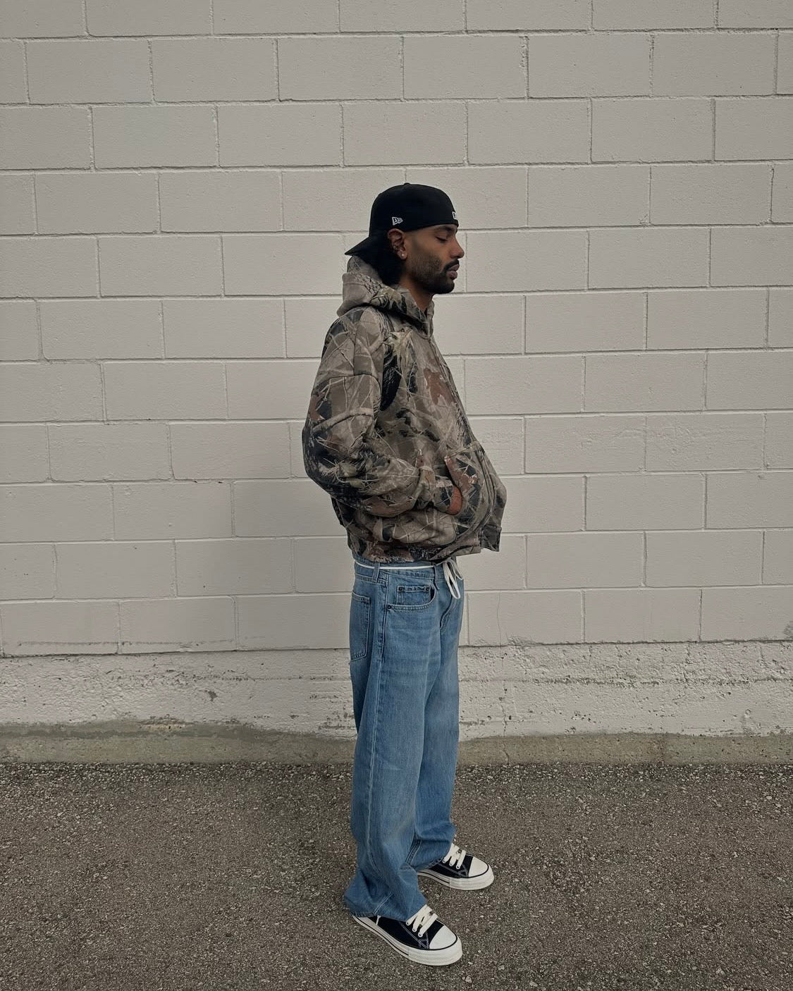 Forest camo hoodie worn in profile against a concrete block wall